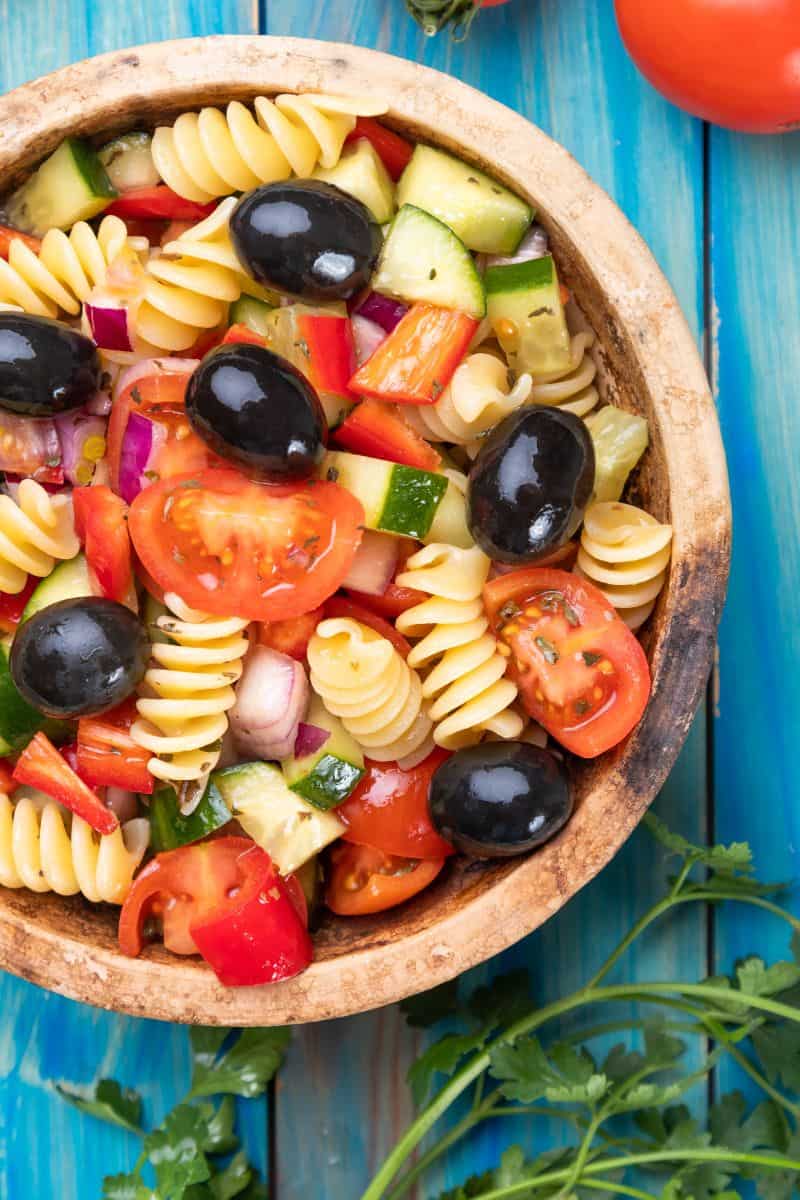 pasta salad in a wooden bowl with rotini pasta, cucumbers, tomatoes, red onions, peppers, and olives.