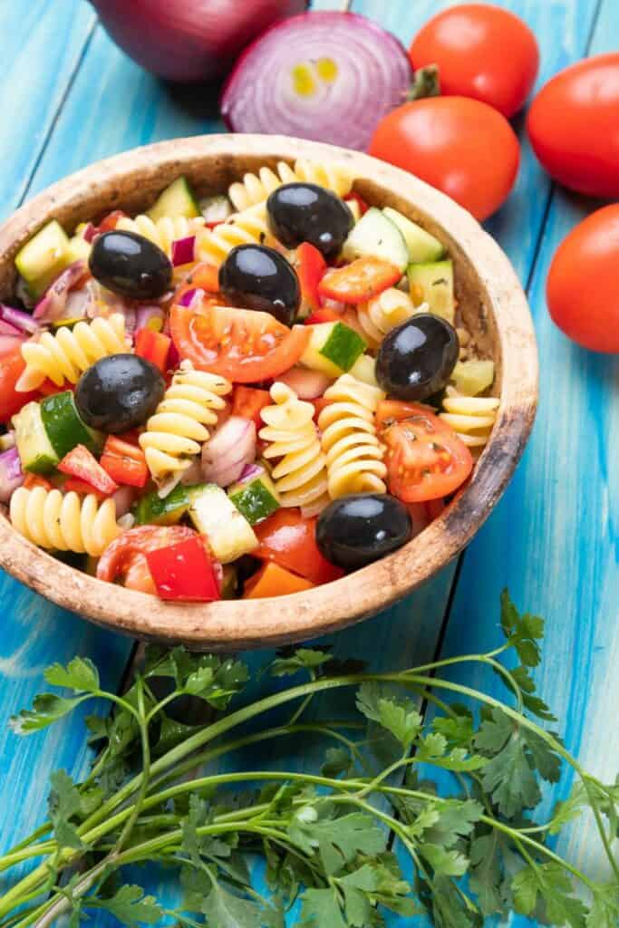 pasta salad in a wooden bowl with red onions, tomatoes, and parsley around it